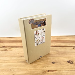 May include: A beige photo album with a label that reads "300 Easy Slip-In Pockets Photo Album" and features a vintage illustration of people playing baseball. The album has a red, white, and blue star on the front.