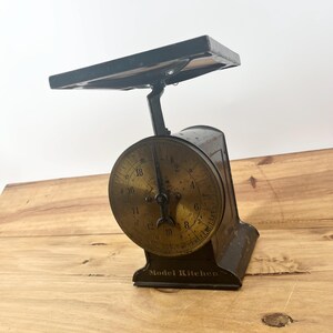May include: A vintage Model Kitchen scale featuring a dark metal frame and a circular brass-toned dial with black numerical markings. The scale has a flat rectangular weighing platform at the top and a single black metal pointer centered on the dial.