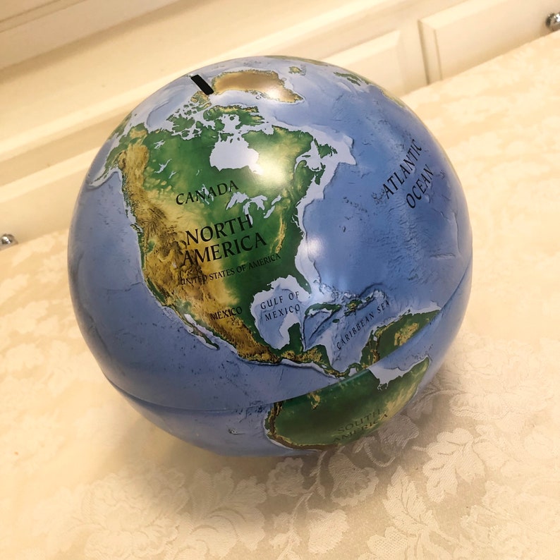 May include: A blue and green globe piggy bank with a slot on the top. The globe shows the continents of North and South America, as well as the Atlantic Ocean. The text "CANADA", "NORTH AMERICA", "UNITED STATES OF AMERICA", "MEXICO", "GULF OF MEXICO", "CARIBBEAN SEA", and "ATLANTIC OCEAN" is printed on the globe.