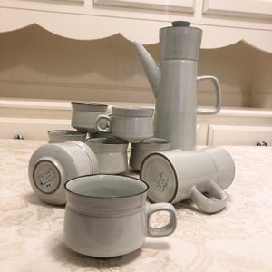 May include: A set of white ceramic coffee mugs and a matching coffee pot. The mugs have a brown rim and the coffee pot has a brown lid. The set is on a white tablecloth.