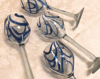 May include: Four clear wine glasses with blue abstract designs. The glasses have long stems and wide, rounded bowls. The blue paint is swirled and streaked across the glass. The glasses are arranged on a patterned, off-white surface.