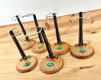 May include: A collection of six doll stands. Each stand features a round, light-brown wooden base with a green and white label. A black metal pole extends upwards, supporting a silver metal hoop. The stands are arranged on a wooden surface.