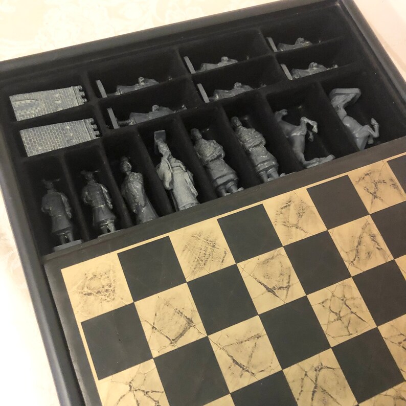 May include: A wooden chessboard with a black and white checkered pattern. The board is open and shows a set of gray chess pieces in the slots above the board. The pieces are arranged in a row and include a king, queen, bishop, knight, rook, and pawn.
