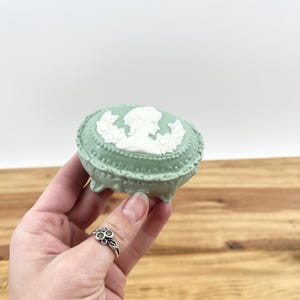 May include: A small, oval-shaped, mint green jewellery box with a white cameo design on the lid. The box has a decorative border and sits on small feet. The cameo features a profile of a woman.