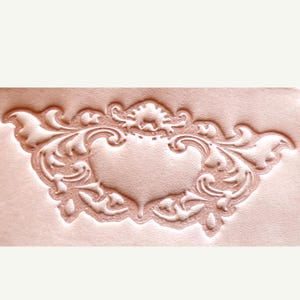 May include: A light beige leather surface featuring a deeply embossed, symmetrical decorative pattern. The design consists of a central shield-like shape surrounded by intricate, flowing scrollwork and leaf-like motifs in a relief style.