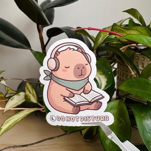 May include: A sticker featuring a cartoon capybara wearing headphones and a green scarf, reading a book. The sticker has the text "DO NOT DISTURB" at the bottom. The sticker is surrounded by green plants.