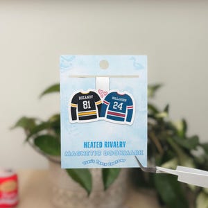 May include: A magnetic bookmark featuring two hockey jerseys with the names "Rozanov" and "Hollander" and jersey numbers 81 and 24. The bookmark has the text "Heated Rivalry Magnetic Bookmark" and the company name "Yuzu's Paper Company".