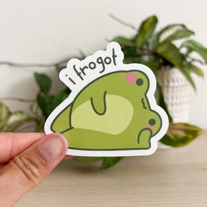 May include: A die-cut sticker featuring a cartoon frog in shades of green with the text "i frogot". The frog has pink cheeks and a light green belly. The sticker is held in a hand.
