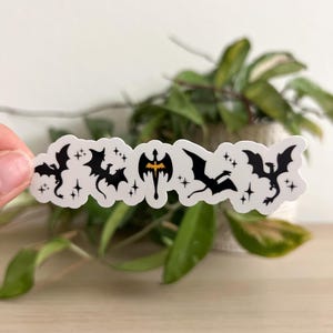 May include: A horizontal white sticker featuring a series of black dragon silhouettes in various flying poses, interspersed with small four-pointed stars. The central dragon is depicted in a symmetrical, upright position with a vibrant orange body.