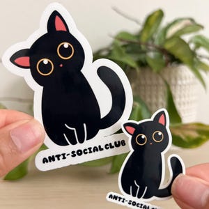 Puede incluir: Dos pegatinas de gato negro con el texto "ANTI-SOCIAL CLUB". Los gatos tienen ojos grandes y redondos, orejas internas rosas y contornos blancos. Una pegatina es m&aacute;s grande que la otra.