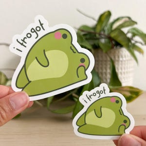 Op de afbeelding: Twee groene kikkerstickers met de tekst "i frogot". De kikkerstickers hebben een lichtgroen lichaam, een donkerdere groene buik en roze wangen. De ene sticker is groter dan de andere.