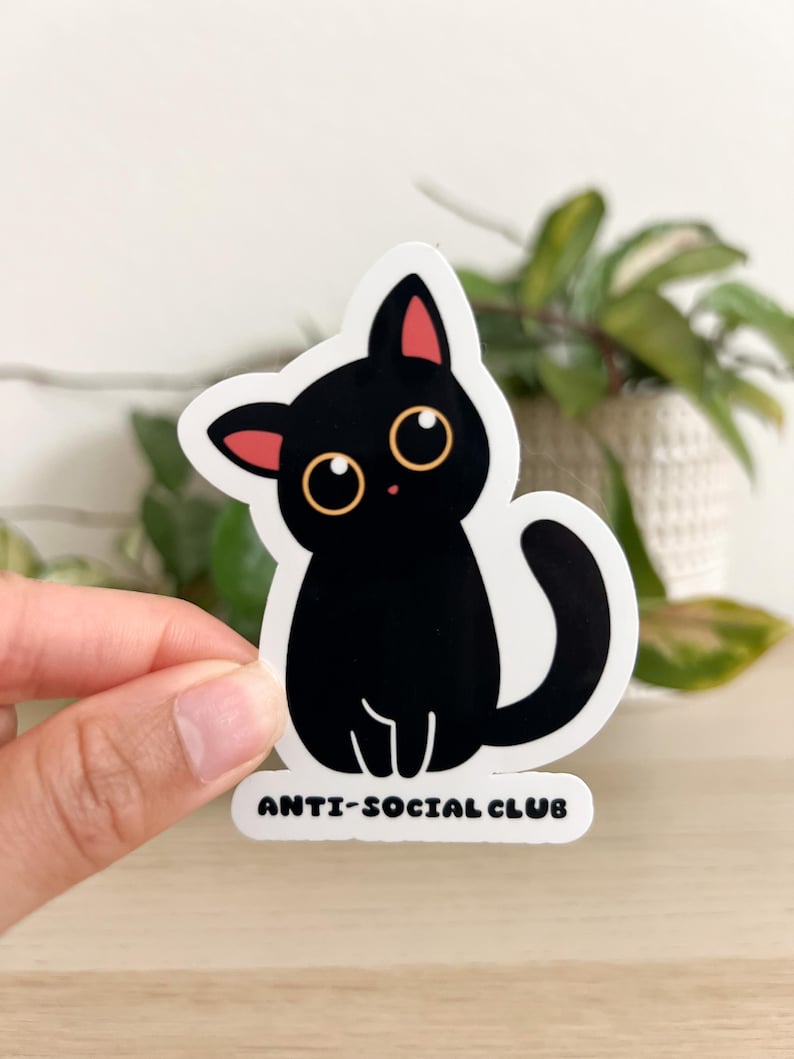 Puede incluir: Una pegatina de gato negro con ojos grandes y redondos y orejas internas rosas. La pegatina tiene el texto "ANTI-SOCIAL CLUB" en la parte inferior. El gato est&aacute; sentado con la cola enroscada. La pegatina est&aacute; sobre un fondo blanco.
