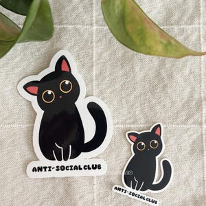 Puede incluir: Dos pegatinas de gato negro con el texto "ANTI-SOCIAL CLUB". Los gatos tienen orejas internas rosas y ojos grandes y redondos. Una pegatina es m&aacute;s grande que la otra. Las pegatinas est&aacute;n sobre una superficie texturizada de color neutro.