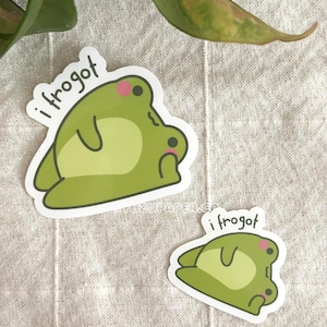 Op de afbeelding: Twee stickers met een cartoon kikker ontwerp in groentinten, met de tekst "i frogot". De kikker heeft een lichtgroen lichaam, een witte buik en roze wangen. De stickers liggen op een neutraal gekleurde stof.