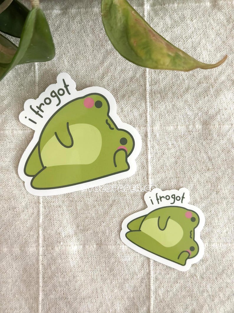 Op de afbeelding: Twee stickers met een cartoon kikker ontwerp in groentinten, met de tekst "i frogot". De kikker heeft een lichtgroen lichaam, een witte buik en roze wangen. De stickers liggen op een neutraal gekleurde stof.
