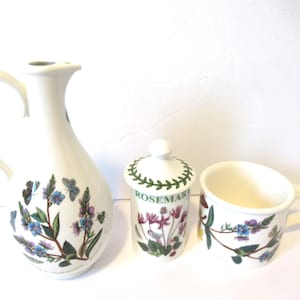 May include: A white ceramic pitcher, a small jar with a lid, and a mug. The pitcher and mug have a handle. All three items are decorated with blue and purple flowers, butterflies, and green leaves.