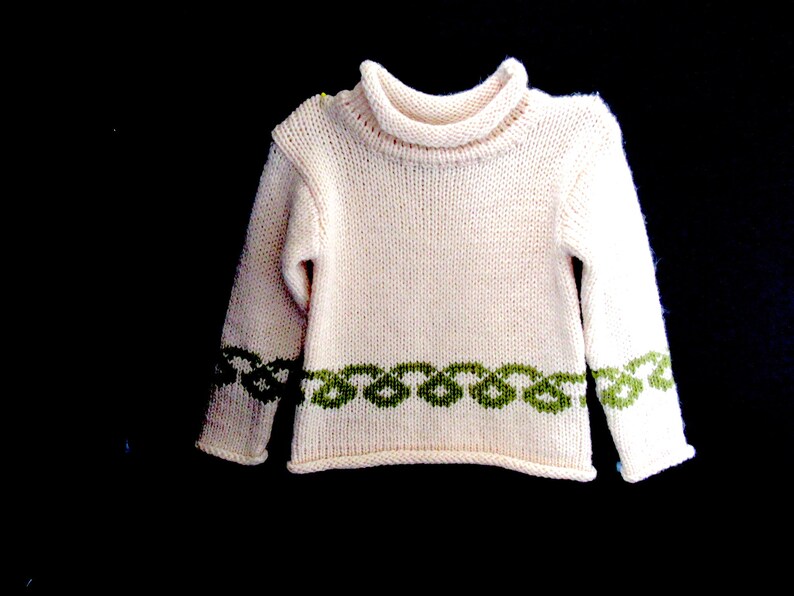 Pu&ograve; includere: Un maglione di lana bianco con un bordo verde intorno al collo e ai polsini. Il maglione ha un collo alto.
