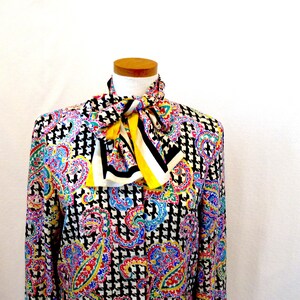 Pu&ograve; includere: Una camicetta a maniche lunghe con un colorato motivo paisley nei toni del blu, rosso, giallo e viola. La camicetta presenta un motivo a pied-de-poule bianco e nero e un papillon a righe gialle, nere e bianche.
