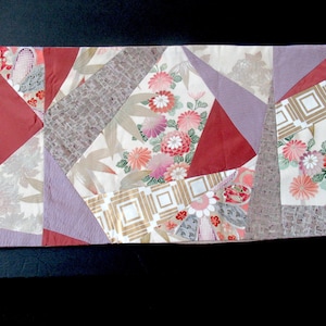 May include: A patchwork textile featuring a variety of fabrics in a geometric design. The colors include burgundy, lavender, cream, and gold. Floral and geometric patterns are visible. The textile is likely a table runner or decorative piece.
