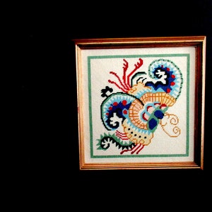 May include: A framed cross-stitch embroidery featuring a colorful, abstract design with blue, yellow, red, and green hues. The design resembles a stylized floral or paisley pattern.