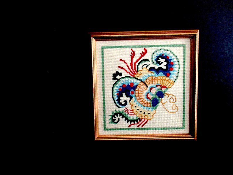 May include: A framed cross-stitch embroidery featuring a colorful, abstract design with blue, yellow, red, and green hues. The design resembles a stylized floral or paisley pattern.