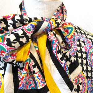 Pu&ograve; includere: Una sciarpa di seta colorata con un motivo a pied-de-poule in bianco e nero, accentuata da disegni paisley in rosa, blu, verde e giallo. La sciarpa &egrave; annodata, mostrando i suoi colori vivaci e i motivi intricati.
