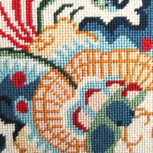 May include: Close-up of a colorful needlepoint design featuring a floral motif with red, blue, green, and yellow threads. The design is stitched on a white canvas.