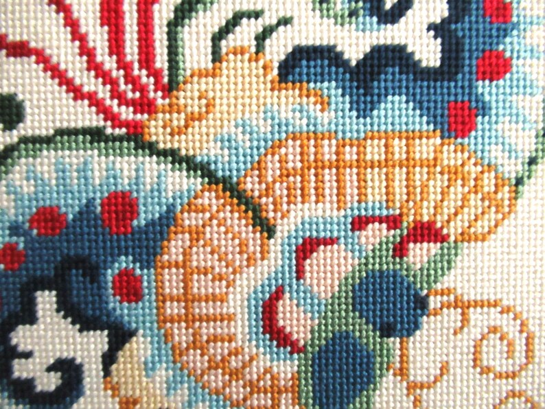 May include: Close-up of a colorful needlepoint design featuring a floral motif with red, blue, green, and yellow threads. The design is stitched on a white canvas.