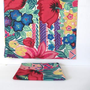 May include: A rectangular fabric piece featuring a vibrant floral pattern with large red poppies, blue blossoms, yellow flowers, and green foliage on a white background. A smaller, folded matching fabric square is positioned in front of the larger piece.