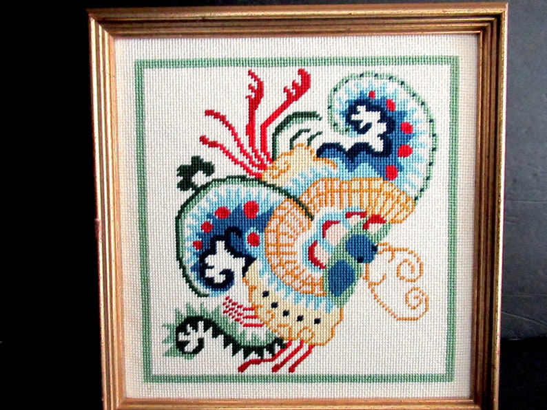 May include: A framed cross-stitch embroidery depicting a colorful, stylized dragon with blue, red, yellow, and green accents.
