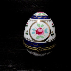 May include: A white porcelain egg-shaped trinket box with a blue and gold floral design. The lid features a pink rose with green leaves and gold accents.