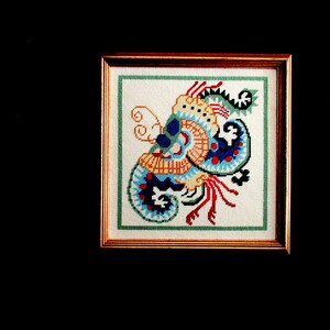 May include: A framed cross-stitch embroidery featuring a colorful, abstract design with blue, red, yellow, and green hues. The design resembles a stylized floral or paisley pattern.
