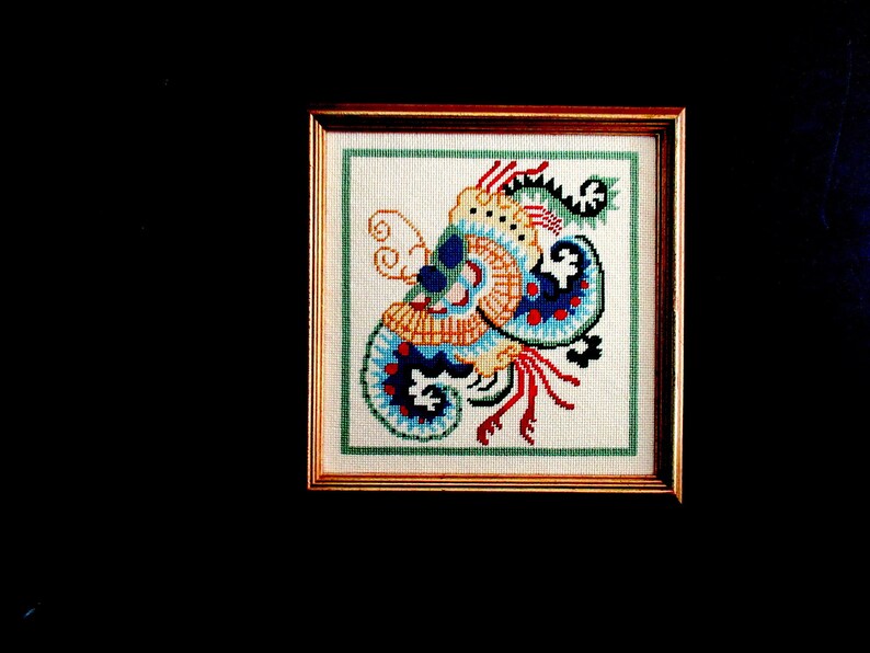 May include: A framed cross-stitch embroidery featuring a colorful, abstract design with blue, red, yellow, and green hues. The design resembles a stylized floral or paisley pattern.