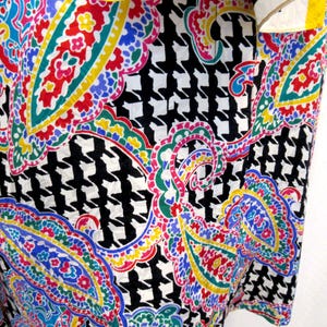 Pu&ograve; includere: Primo piano di un tessuto con un vivace motivo paisley e pied-de-poule. Il design paisley presenta accenti rossi, blu, gialli e verdi su sfondo bianco. Il motivo pied-de-poule &egrave; bianco e nero.