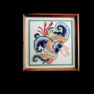 May include: A framed cross-stitch embroidery featuring a colorful abstract design with blue, red, yellow, and green hues. The design resembles a stylized floral or animal motif.