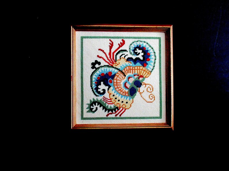 May include: A framed cross-stitch embroidery featuring a colorful abstract design with blue, red, yellow, and green hues. The design resembles a stylized floral or animal motif.