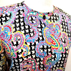 Pu&ograve; includere: Una camicetta colorata con un motivo a pied-de-poule bianco e nero, sovrapposta a vivaci disegni paisley nei toni del rosso, blu, giallo e verde. La camicetta ha uno scollo tondo e un singolo bottone nero.