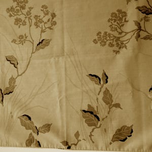 May include: A light tan fabric featuring a repeating pattern of dark brown botanical illustrations, including leafy branches, small clusters of flowers, and perched birds. The design is printed in a monochromatic style across the textile surface.