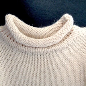 Pu&ograve; includere: Primo piano di un maglione di lana color crema con collo alto ripiegato.