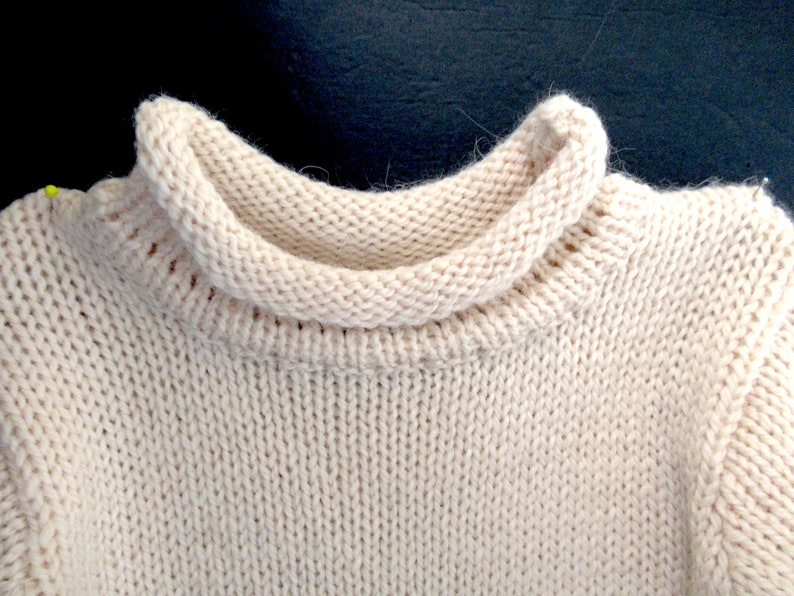 Pu&ograve; includere: Primo piano di un maglione di lana color crema con collo alto ripiegato.