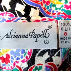 Pu&ograve; includere: Primo piano di un tessuto di seta colorato con un motivo paisley in rosso, rosa, blu, verde e giallo. L'etichetta recita "Adrianna Papell" e "100% Seta, Taglia 8, Made in Korea."
