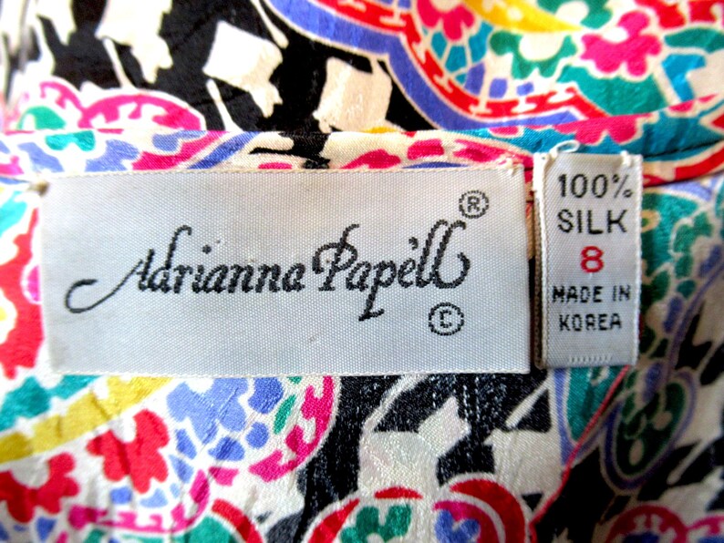 Pu&ograve; includere: Primo piano di un tessuto di seta colorato con un motivo paisley in rosso, rosa, blu, verde e giallo. L'etichetta recita "Adrianna Papell" e "100% Seta, Taglia 8, Made in Korea."