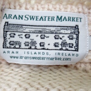 Pu&ograve; includere: Etichetta di maglione Aran bianco con il testo "Aran Sweater Market" e un'illustrazione in bianco e nero di un edificio. L'etichetta include anche il testo "Aran Islands, Ireland" e "www.aransweatermarket.com". L'etichetta ha una piccola etichetta bianca attaccata con la lettera "S" stampata su di essa.