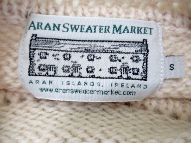 Pu&ograve; includere: Etichetta di maglione Aran bianco con il testo "Aran Sweater Market" e un'illustrazione in bianco e nero di un edificio. L'etichetta include anche il testo "Aran Islands, Ireland" e "www.aransweatermarket.com". L'etichetta ha una piccola etichetta bianca attaccata con la lettera "S" stampata su di essa.
