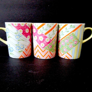 May include: Three ceramic mugs with colorful patchwork designs. Each mug has a different pattern featuring geometric shapes, flowers, and stripes. The mugs have yellow handles.