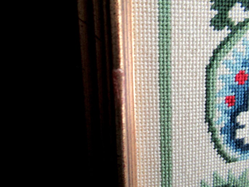 May include: Close-up of a framed needlepoint piece with a floral design. The design features blue, green, and red flowers on a white background.