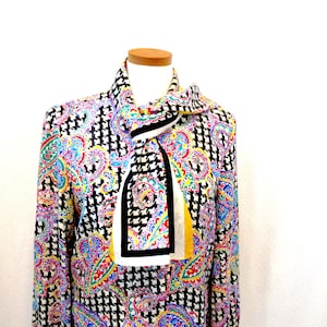 Pu&ograve; includere: Un completo camicetta e sciarpa fantasia. La camicetta presenta un motivo pied-de-poule bianco e nero con dettagli paisley colorati in rosa, blu, giallo e verde. La sciarpa ha un design geometrico in bianco e nero con una striscia gialla.