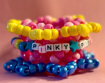 May include: A stack of colorful beaded bracelets in shades of pink, yellow, and blue. One bracelet spells out "PINKY PIE" with white letter beads and star accents. The bracelets are stacked on a pink background.
