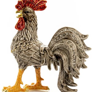 Könnte beinhalten: Dekorative Hahnfigur mit rotem Kamm und Kehllappen, grauem Körper und gelben Füßen. Die Schwanzfedern sind mit kleinen, funkelnden Kristallen verziert. Der Hahn steht auf einer weißen Oberfläche.
