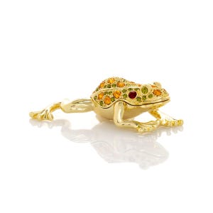 May include: A gold-colored frog figurine with a jeweled back and eyes. The frog's back is covered in orange, yellow, and green gemstones. The eyes are red and green. The frog is lying on a white surface.